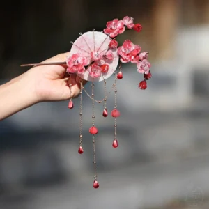 Handmade plum blossom Chinese hair stick with miniature oil paper umbrella and layered red tassels held against a traditional outdoor backdrop