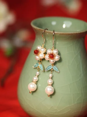 Plum blossom palace Chinese earrings displayed on a green ceramic vase with white flowers red crystal centers blue enamel leaves and pearl drop pendants