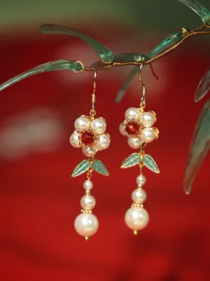 Gold tone plum blossom Hanfu earrings hanging from a green branch against red fabric with layered pearl dangles and blue enamel leaves