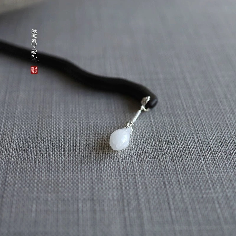 Top-down view of a polished black ebony Chinese hair pin with silver link detail and round white jade pendant on grey linen fabric