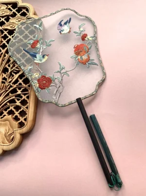 Embroidered pomegranate and songbird Chinese palace hand fan with translucent fabric, peonies, flowering branches, scalloped decorative border, dark bamboo handle, and green handle cover displayed on a light pink tabletop