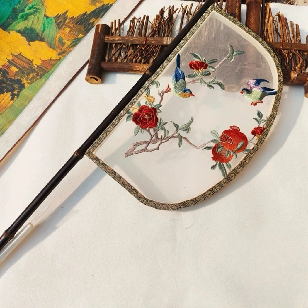 Oblique top view of an embroidered knife-shaped Chinese hand fan with a sheer silk-style fan surface, dark bamboo handle, pomegranate and songbird motifs, traditional brocade border, bamboo crafts, and Chinese landscape decor
