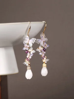 Close-up pair of purple butterfly drop earrings with amethyst-style bead clusters, white teardrop charms and gold-tone details on a dark background