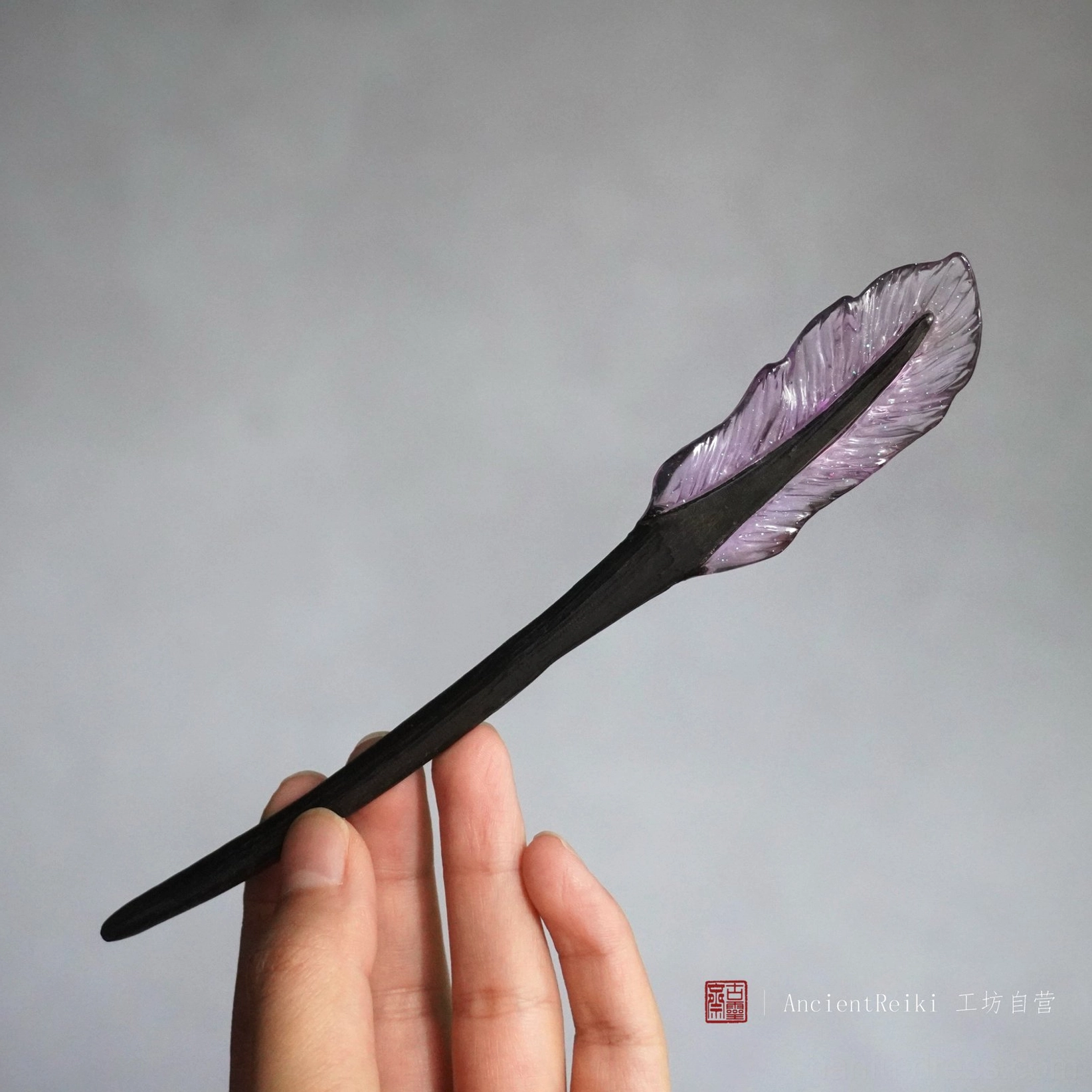 Purple feather Chinese hair stick held in hand, showing the polished black wooden shaft and translucent feather ornament