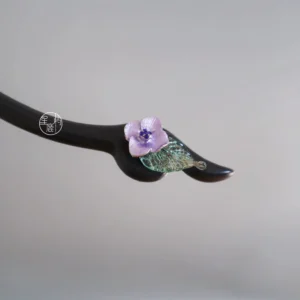 Purple flower Chinese hair stick product close-up on grey background with polished black wooden pin and textured green leaf decoration