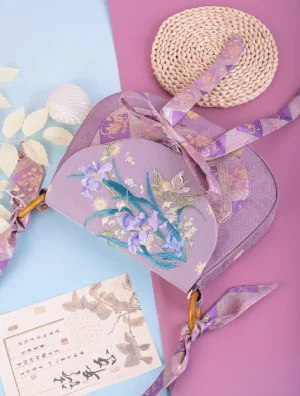 Angled view of purple iris embroidered Chinese crossbody bag showing bamboo style buckle, multicolor floral embroidery, lavender jacquard strap and decorative seashell props
