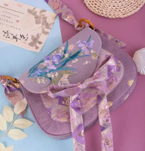 Purple iris embroidered Chinese style crossbody bag in mauve lavender fabric with floral flap embroidery, gold jacquard bow strap, calligraphy postcards and rattan styling props