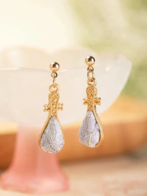 Purple pipa Chinese drop earrings with carved gold-toned metal details displayed on a pink glass cup and wooden tray, elegant jewelry for Hanfu Qipao and Cheongsam styling
