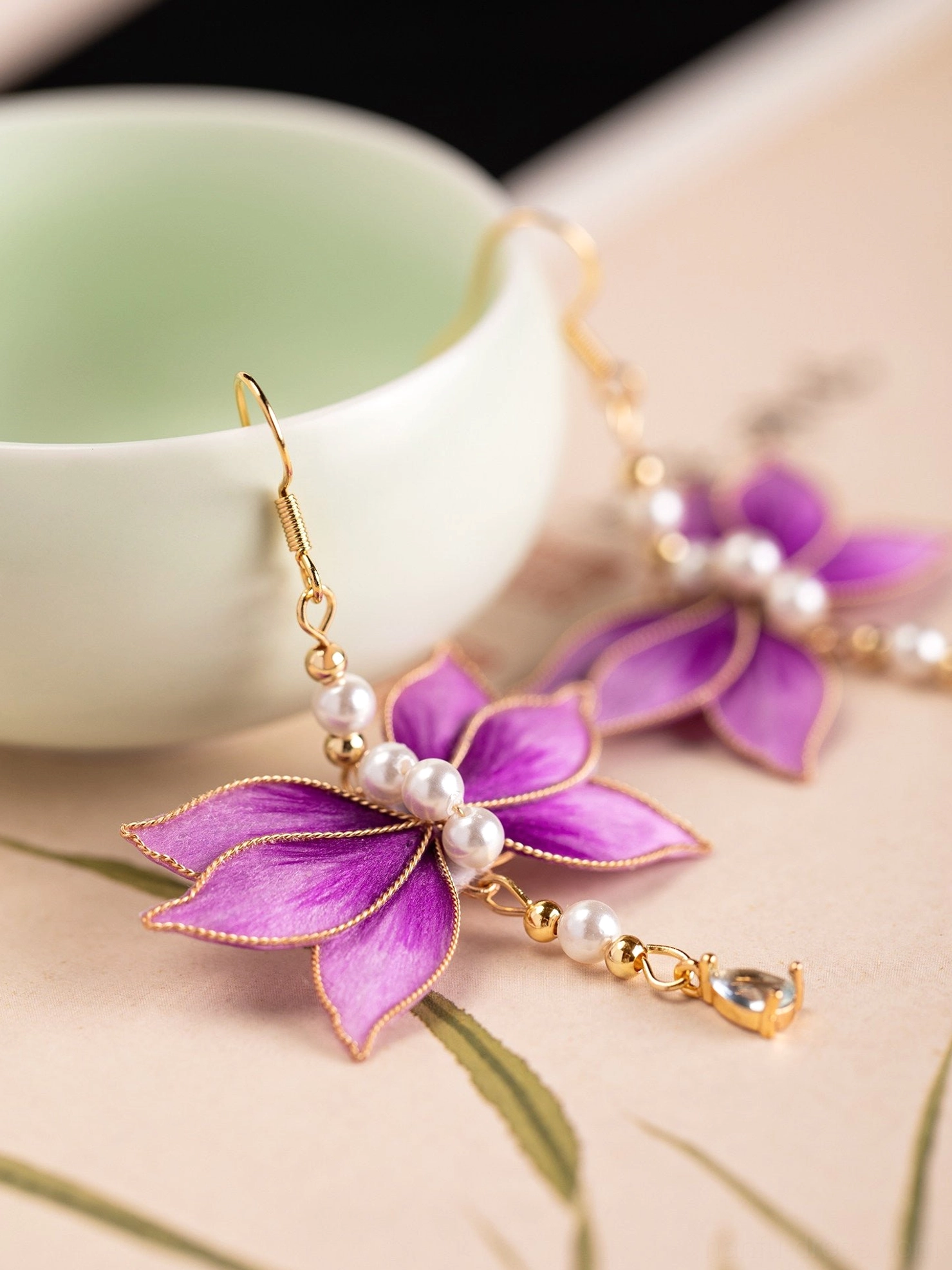 Purple velvet lotus Hanfu earrings with gold edged petals pearl details and dangling charms beside a traditional teacup