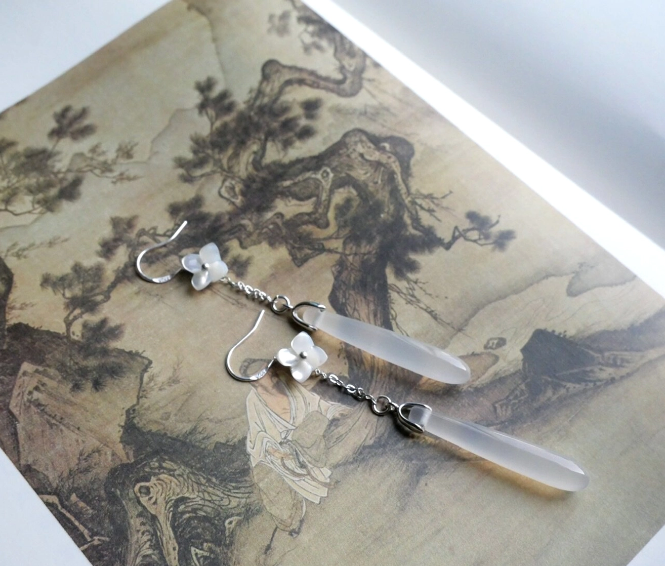 Pair of Rain Alley floral Chinese hook earrings on traditional landscape painting, featuring white flower charms and long translucent teardrop drops