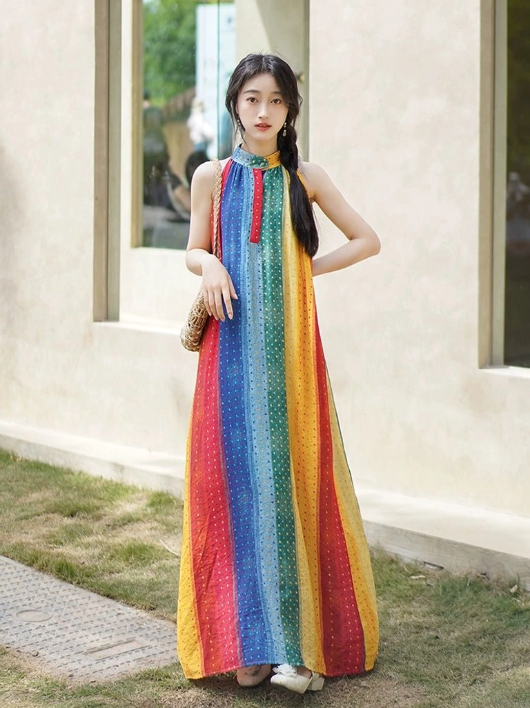 Outdoor side view of a sleeveless rainbow modern Hanfu-inspired maxi dress with stand halter collar, vertical striped polka dot pattern, loose floor-length silhouette, and woven straw bag