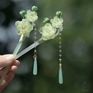 Green Chinese hair stick with translucent flower charms, crystal bead tassels, and jade-inspired pendants held against blurred outdoor foliage