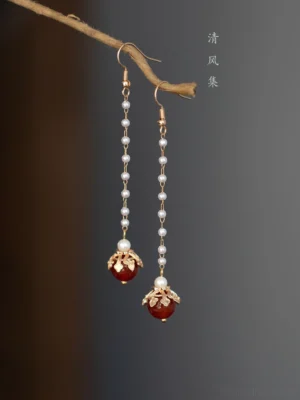 Long red agate Chinese drop earrings with gold floral setting and faux pearl chain displayed on a natural twig against a warm brown background