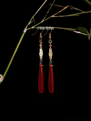 Red agate Hanfu drop earrings with gold Chinese fretwork, red bead accents, and teardrop pendants displayed with green bamboo on a black background