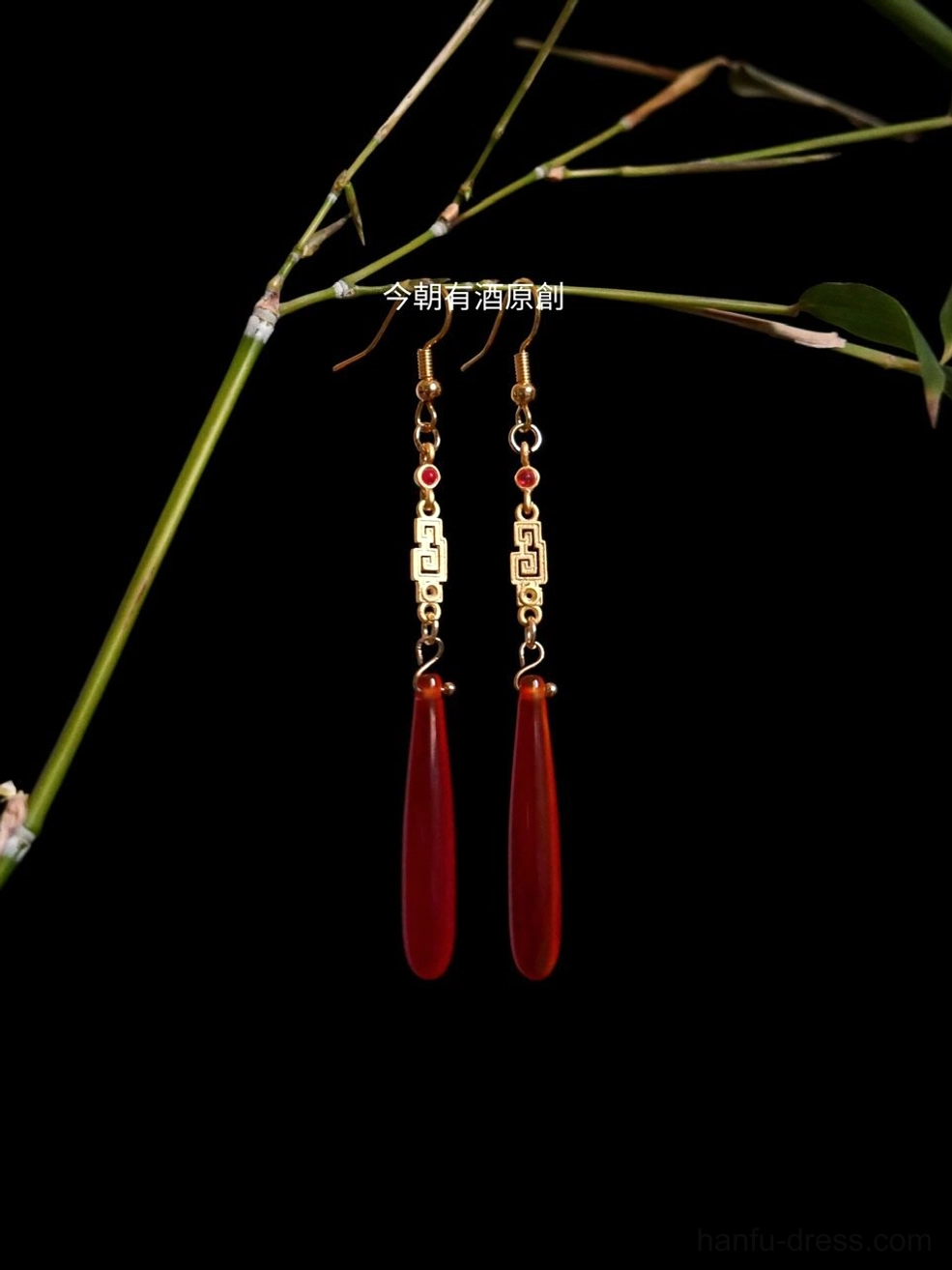 Red agate Hanfu drop earrings with gold Chinese fretwork, red bead accents, and teardrop pendants displayed with green bamboo on a black background