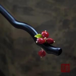 Three-quarter close-up of a red bean Chinese hair stick with a wavy black wooden shaft, red berry clusters, green leaves, golden bead accents, and a dangling red pendant