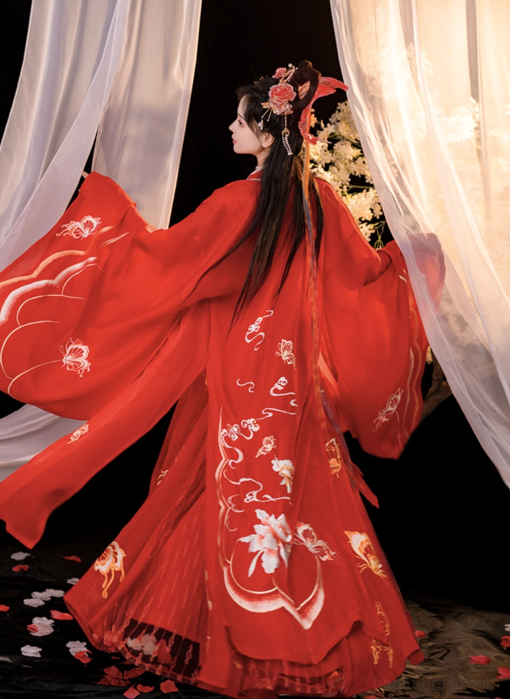 Back three-quarter view of a red Chinese bridal hanfu with intricate golden floral and butterfly embroidery, oversized wide sleeves, long train, and traditional Tang-inspired silhouette
