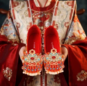 Front view of red Chinese bridal Hanfu shoes with ornate floral toe decorations, gemstone beads, and dangling pearl tassels held with an embroidered Xiuhe wedding outfit