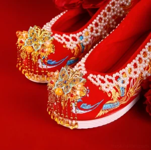 Close-up overhead view of embroidered red Chinese wedding Hanfu shoes featuring phoenix motifs, pearl beaded edging, golden butterfly tassels, and thick white wedge-style soles