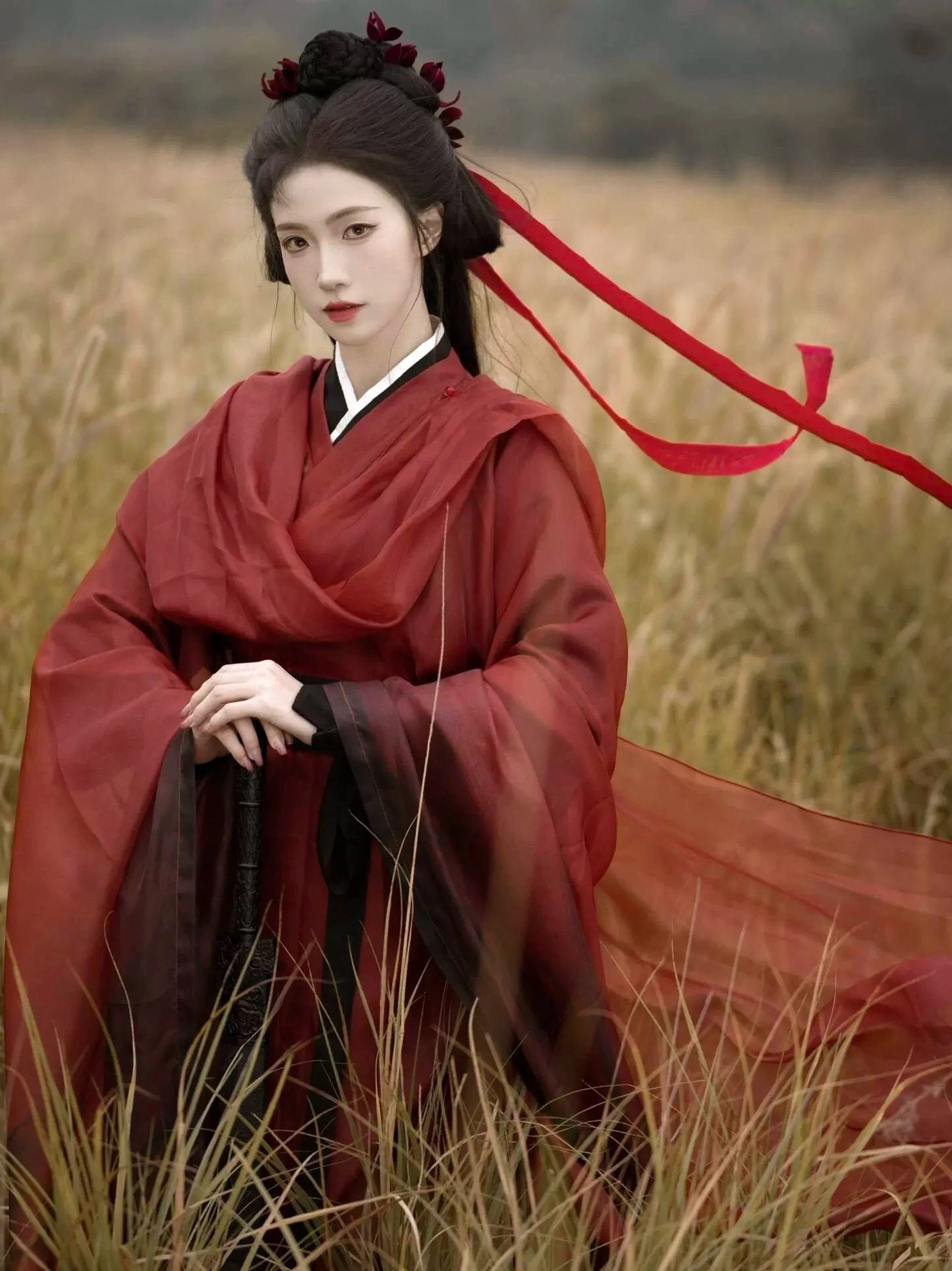 Red chivalrous Hanfu front detail with traditional cross collar, long red hair ribbons, floral hair ornaments, and a flowing robe train beside a sword