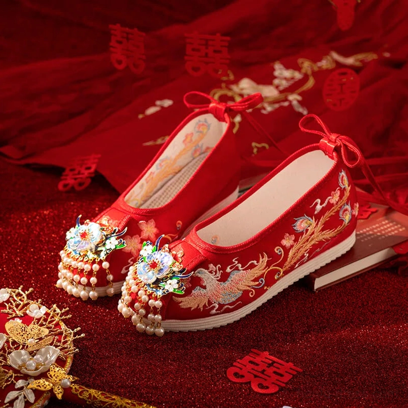 Red dragon and phoenix embroidered Chinese bridal Hanfu shoes with gold floral bead tassel toe decorations on a traditional double happiness wedding backdrop
