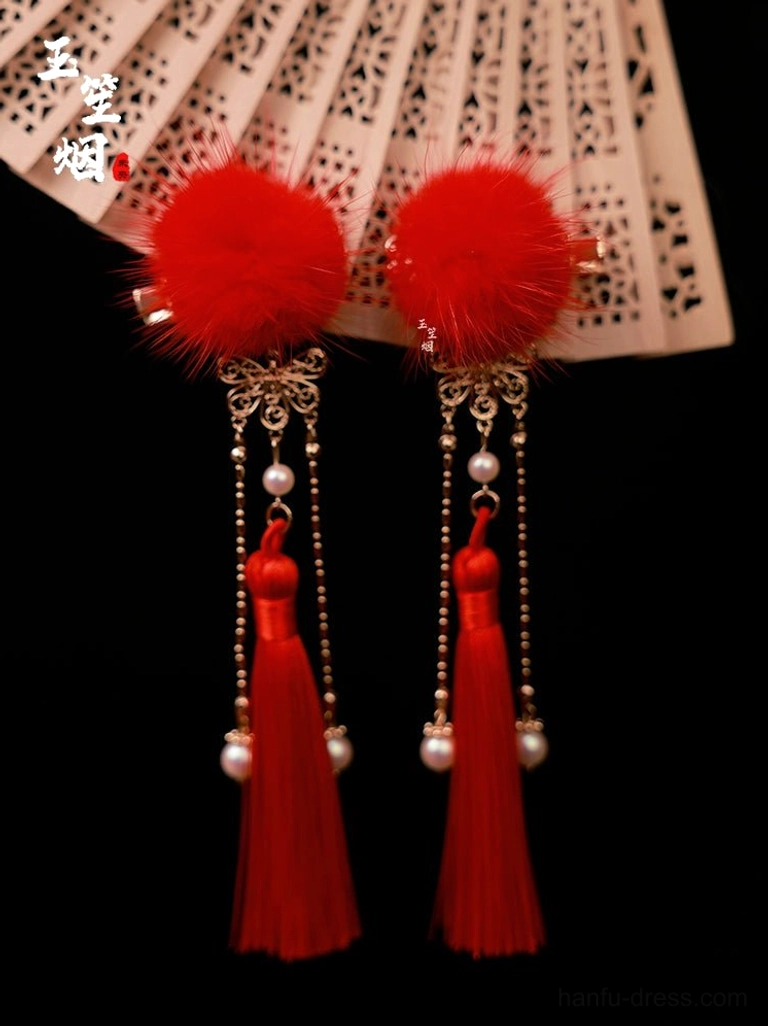 Close-up of red fluffy furball hair pins with golden carved bases pearl bead chains and long red silk tassels beside a Chinese folding fan