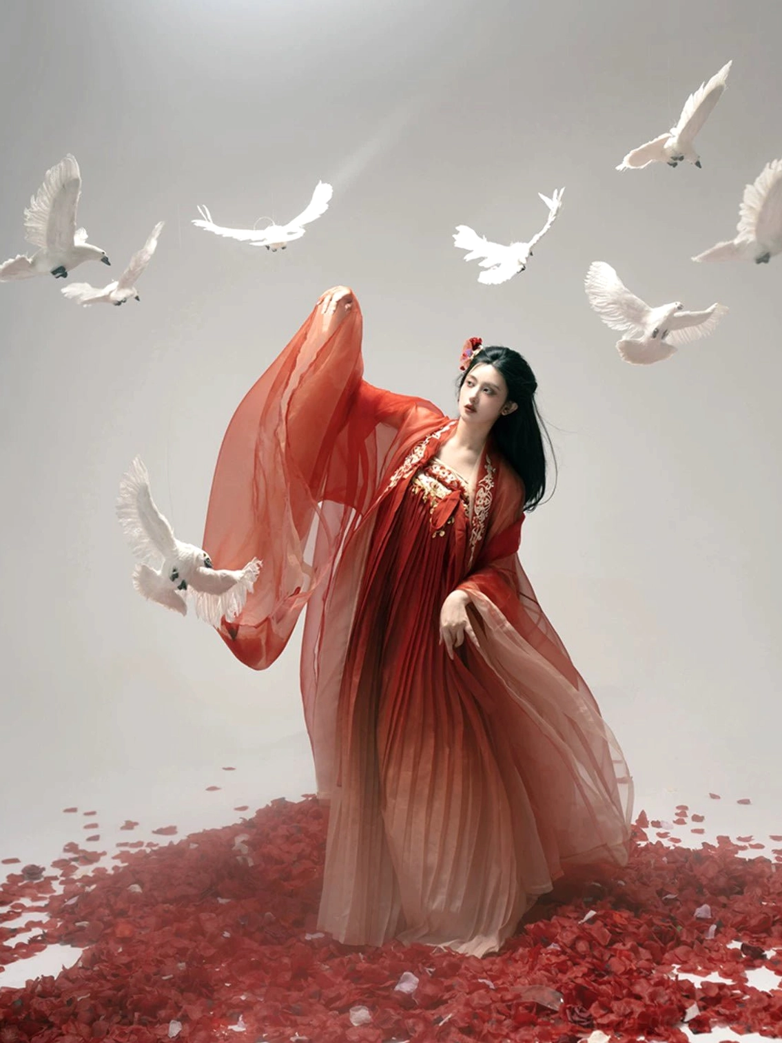 Three-quarter side view of a complete red gradient Tang Dynasty Qixiong Ruqun hanfu set with wide flowing sleeves, pleated chiffon skirt, elevated waistline, rose petals, and white doves