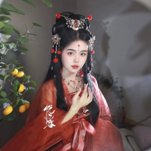 Female model wearing a red traditional Hanfu outfit with Chinese hair ornaments and a warm wine red Chinese necklace styled against a softly lit foliage background