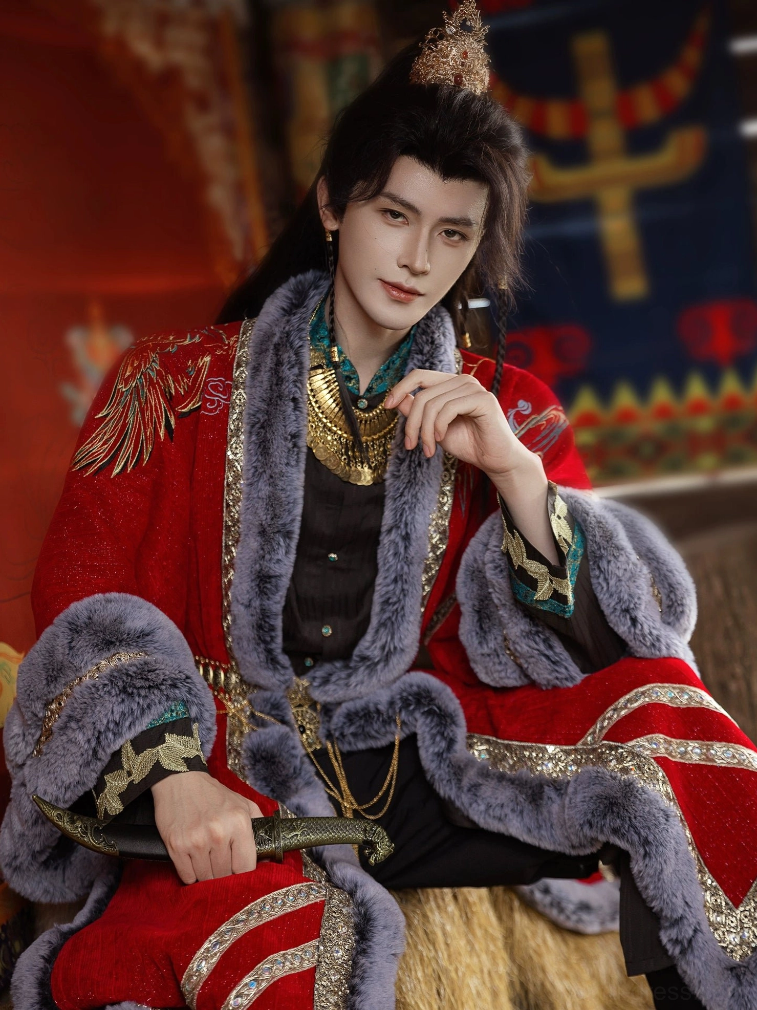 Close-up of a male model wearing a red hanfu overcoat with thick grey faux fur trim, ornate gold neck jewelry, and a decorative miniature sword