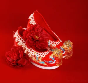Pair of red Chinese bridal Hanfu shoes with blue and gold phoenix embroidery, pearl trim, golden tassel toe ornaments, and layered white rubber soles on a red wedding background