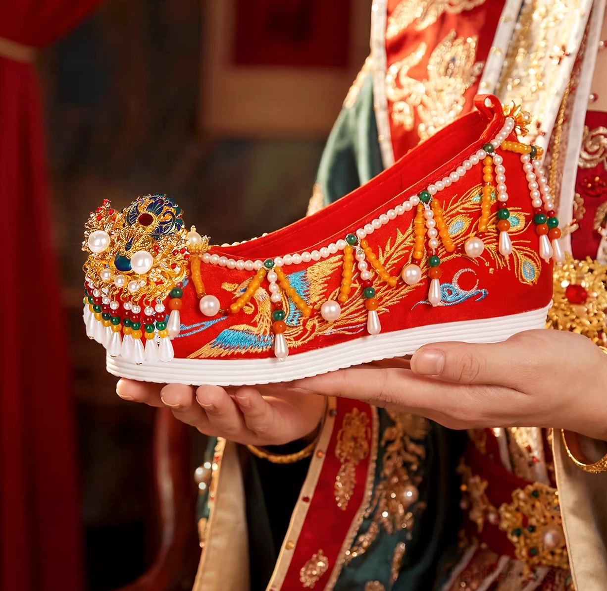 Side view of red Chinese wedding Hanfu shoe with gold phoenix and cloud embroidery, pearl beads, gemstone decorations, tassels, and curved toe