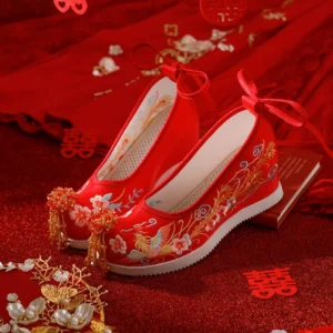 Red phoenix embroidered Chinese bridal Hanfu shoes with beaded tassel toe ornaments, ribbon ties, and white wedge soles displayed on a traditional double happiness wedding backdrop
