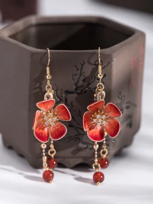 Side view of red plum blossom Hanfu earrings with gold-tone hardware chain tassels and red bead pendants beside a traditional dark ceramic vessel