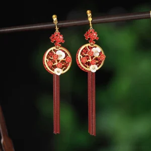 Red plum blossom Chinese hook earrings with gold-edged openwork pendants, red enamel flowers, mother-of-pearl floral accents, and long cascading tassels displayed against a natural green background