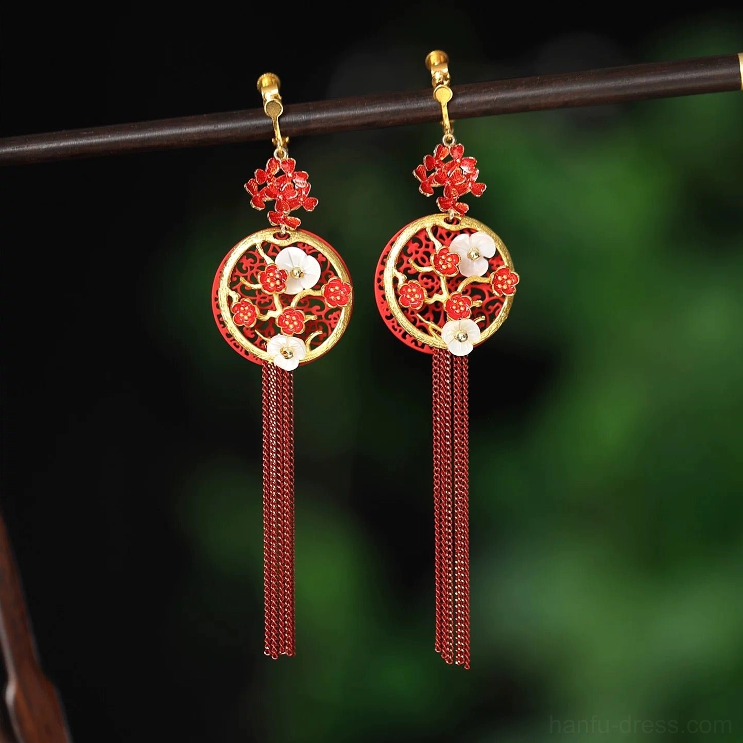 Red plum blossom Chinese hook earrings with gold-edged openwork pendants, red enamel flowers, mother-of-pearl floral accents, and long cascading tassels displayed against a natural green background