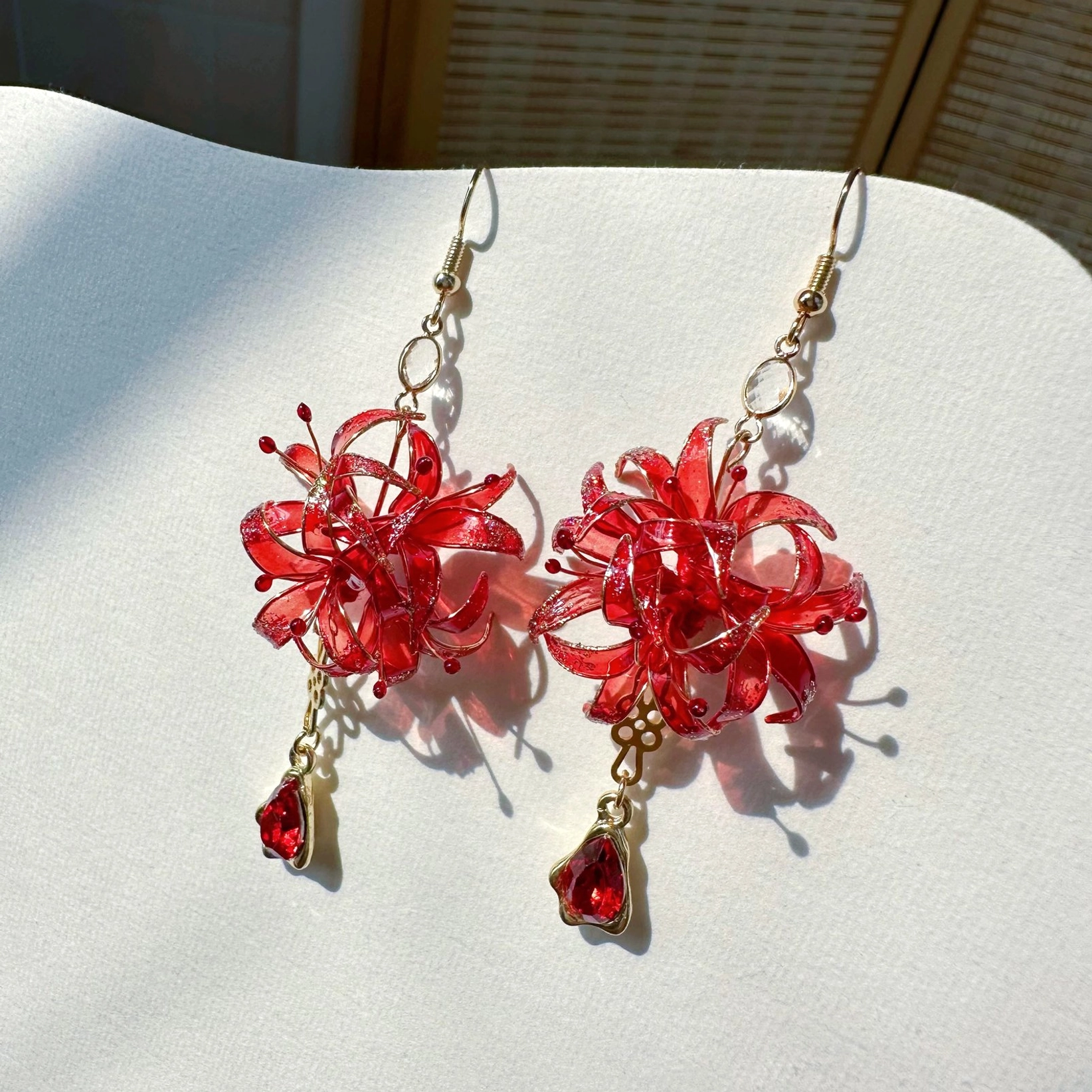 Matching red resin spider lily earrings with fine floral stamens, gold-tone hardware, and teardrop red gemstone-style pendants arranged on an off-white jewelry display board