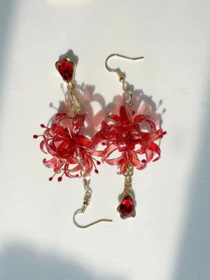 Close-up red spider lily Chinese earrings with detailed flower stamens, gold-tone ear hooks, crystal accents, and teardrop red gemstone-style pendants on a light gray background