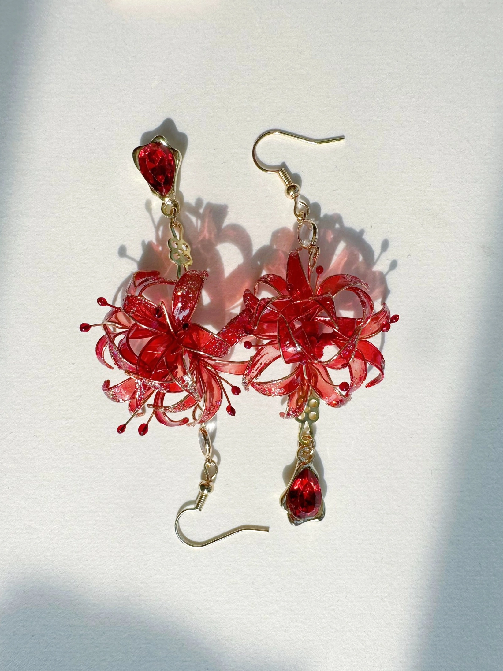 Close-up red spider lily Chinese earrings with detailed flower stamens, gold-tone ear hooks, crystal accents, and teardrop red gemstone-style pendants on a light gray background