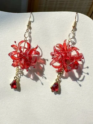 Red spider lily dangle earrings with red flower charms, teardrop red pendants, gold-tone hook wires, and a delicate crystal bead accent displayed on an off-white jewelry board