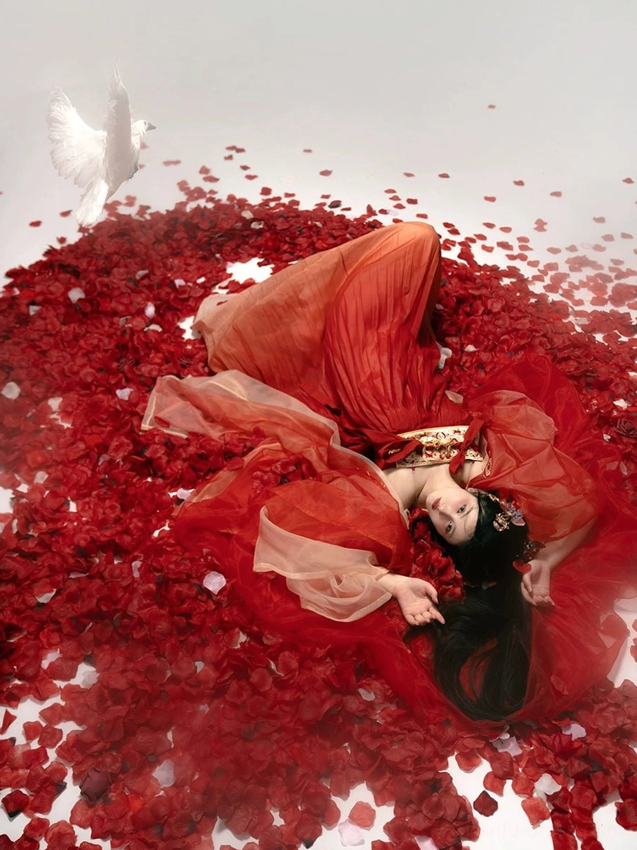 Top-down view of a red Tang Dynasty Qixiong Ruqun hanfu spread across rose petals, showing wide draped sleeves, full pleated skirt, golden chest ornament, and flowing chiffon layers