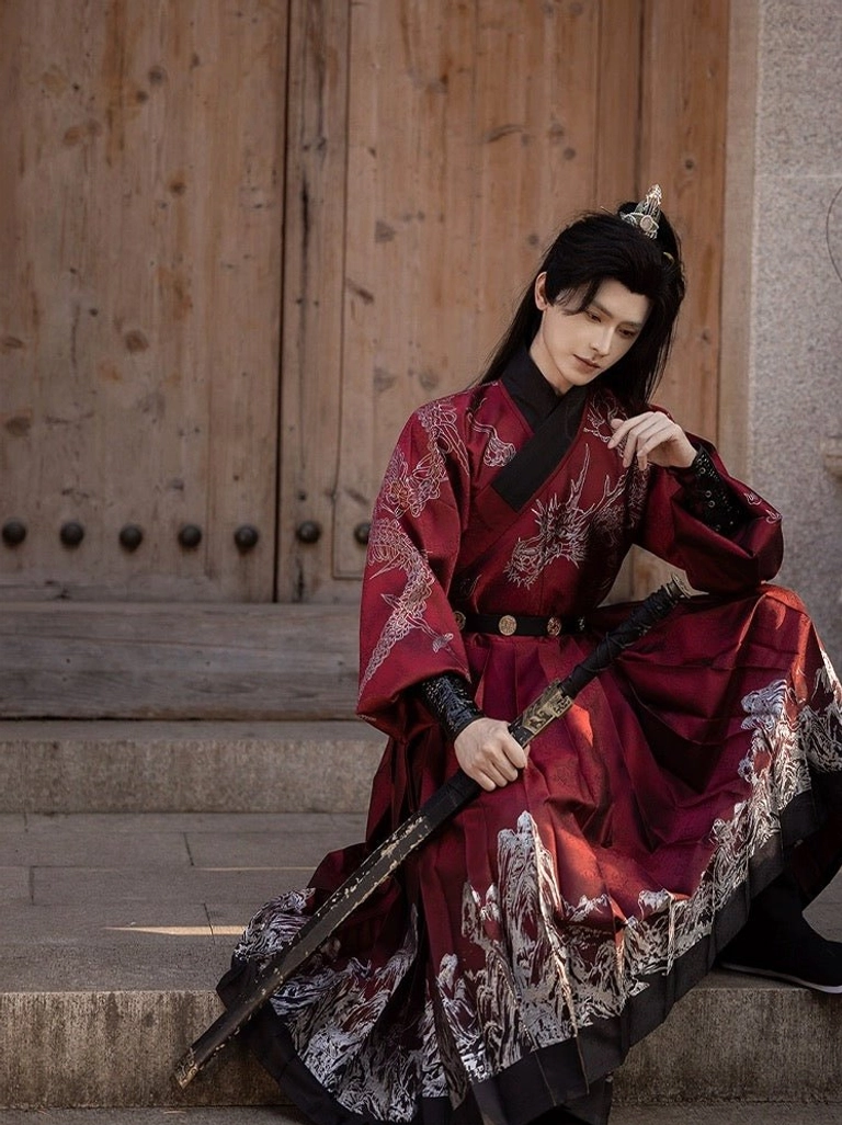 Male model seated on stone steps wearing a red Tie Li Hanfu robe with black wrist guards, matching waist belt, cross collar, and traditional sword