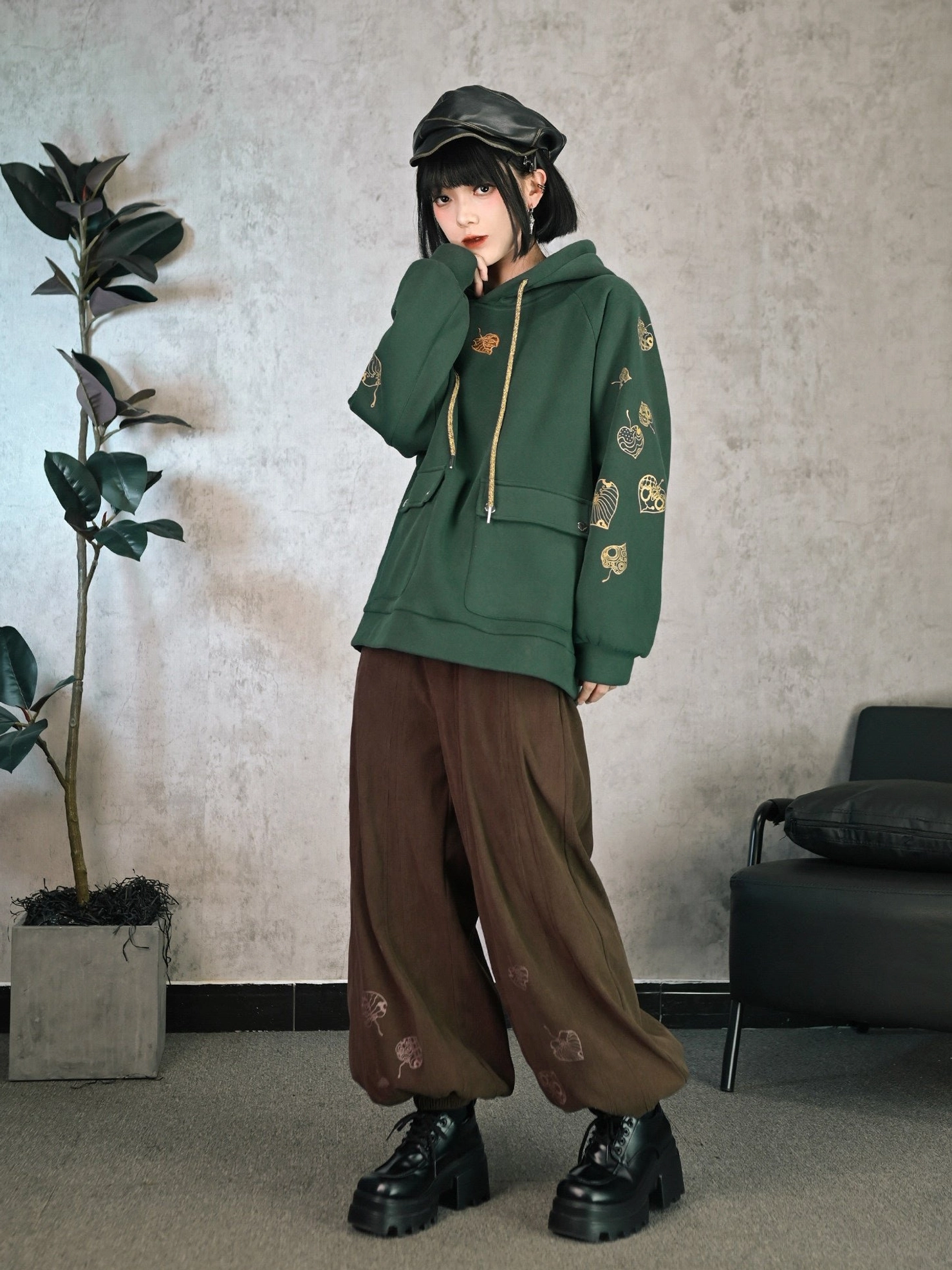 Side full body view of dark green Modern Hanfu hooded fleece sweatshirt with golden botanical embroidery, beige drawstrings, patch pockets, and brown wide leg pants