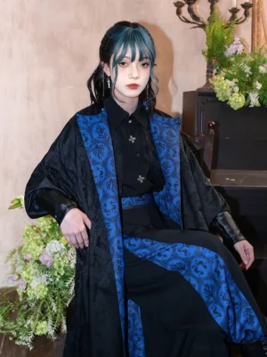 Three-quarter seated view of a reversible modern hanfu long coat showing the blue vintage printed side, loose batwing sleeves, leather-look cuffs, and relaxed Chinese-style silhouette