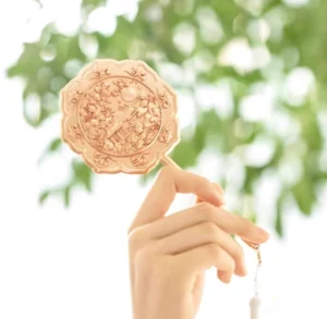 Rose gold Chinese hand mirror held outdoors in natural light, featuring vintage embossed moon palace patterns, floral details, slender handle, and white tassel pendant