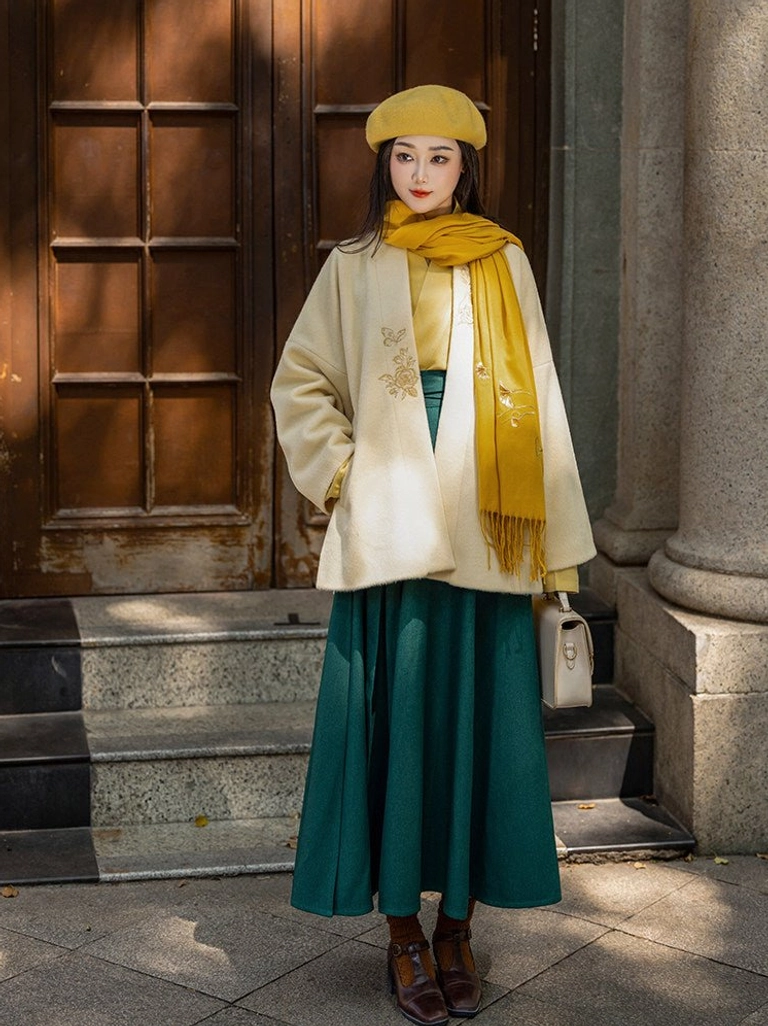 Full-body view of a modern Song Dynasty Hanfu autumn winter outfit with cream embroidered jacket, mustard accessories, and flowing dark green wrap skirt outdoors