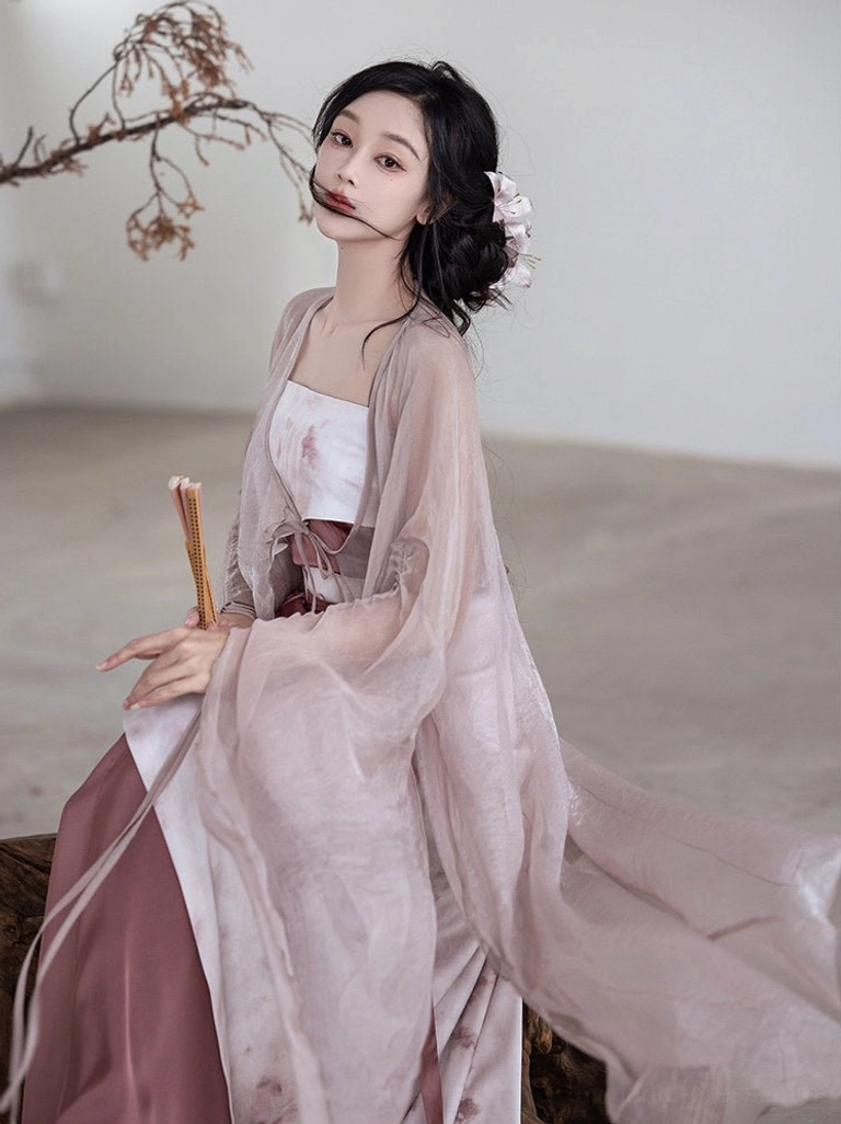Seated side view of modern Song Dynasty Hanfu set with translucent rose outer cardigan, camisole, two tone skirt, floral hair accessory, and folding fan