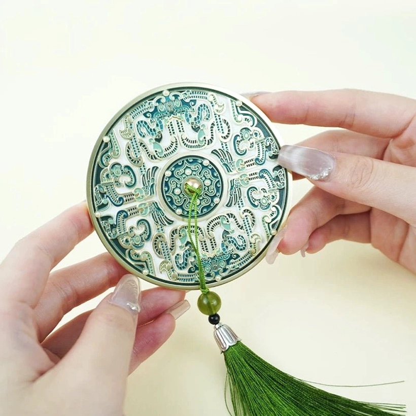 Close-up of a round Chinese pocket mirror with intricate traditional enamel-style patterns, green tassel, and jade-like decorative beads held in both hands