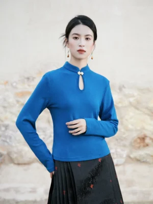 Front three-quarter view of a royal blue Chinese-style knitted top with a classic stand collar, traditional frog fastener, keyhole neckline, and soft fitted silhouette styled with a black pleated skirt
