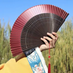 Ruby red traditional Chinese folding hand fan fully opened with dark carved bamboo ribs and red tassel, held with a yellow embroidered Hanfu sleeve outdoors