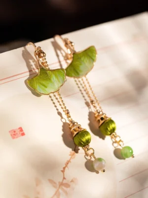 Sage green ginkgo leaf tassel Hanfu earrings with gold tone chains and jade inspired green bead drops displayed on Chinese calligraphy paper